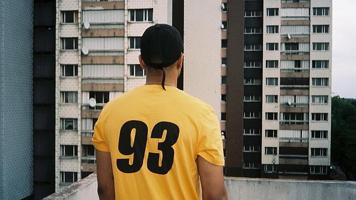 A man in a yellow T-shirt and baseball cap stands on a balcony, his gaze fixed on the wall of the tower block opposite, the number 93 written on his back
