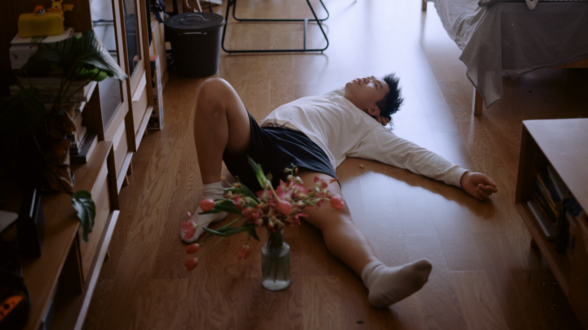 A young man is lying on a wooden floor in a room. One leg is bent, the other stretched out. One arm supports his head, the other is stretched out. At his feet is a vase of flowers. 
