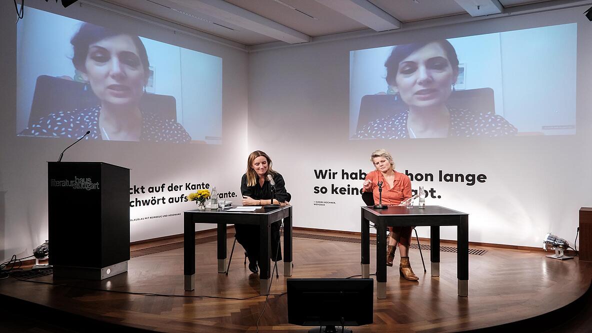 In one corner of a room, two women sit at two black square tables with microphones. The tables are generously placed. On two screens on both visible walls, one can see another woman. The three women are in dialogue with each other. On the left of the picture is a black lectern.