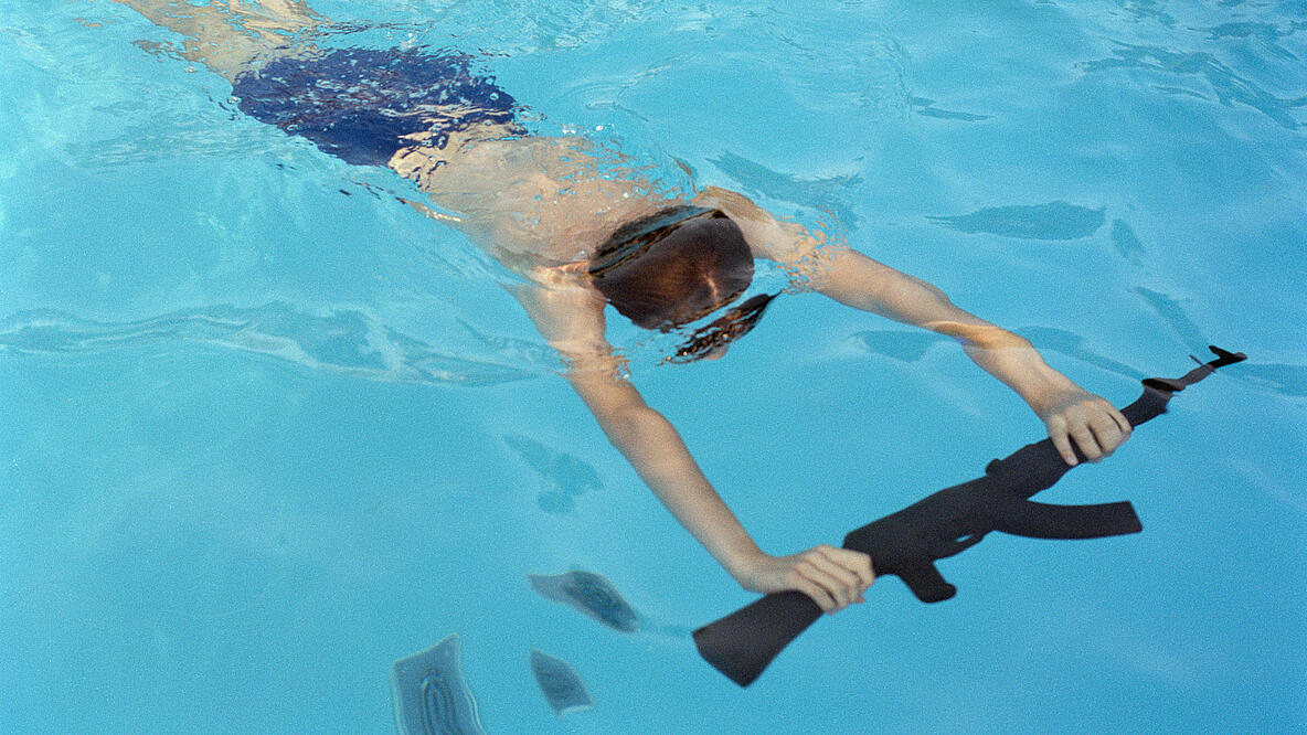 In the clear water of a swimming pool, a boy dives with both arms stretched forward, holding a rifle in both hands.