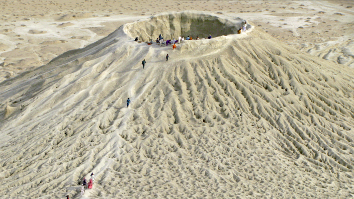 Ein Sandkrater inmitten einer Wüstenlandschaft. Ein Dutzend Menschen klettern den Berg hinauf zum Krater. Ein weiteres Dutzend Menschen steht und sitzt oben am Kraterrand.