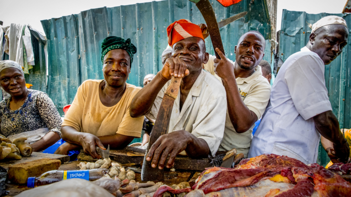 Behind a simple market stall, several traders can be seen looking cheerfully into the camera. Raw meat can be seen on the table, one of the traders leans on his long knife