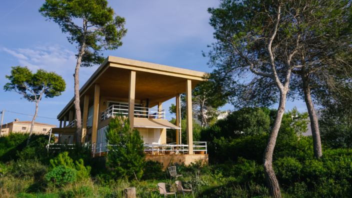 Ein zweigeschossiges sandfarbenes Haus steht in einem grünen Garten. Unterhalb der Terrasse stehen Stühle, ringsum vereinzelt Bäume. 
