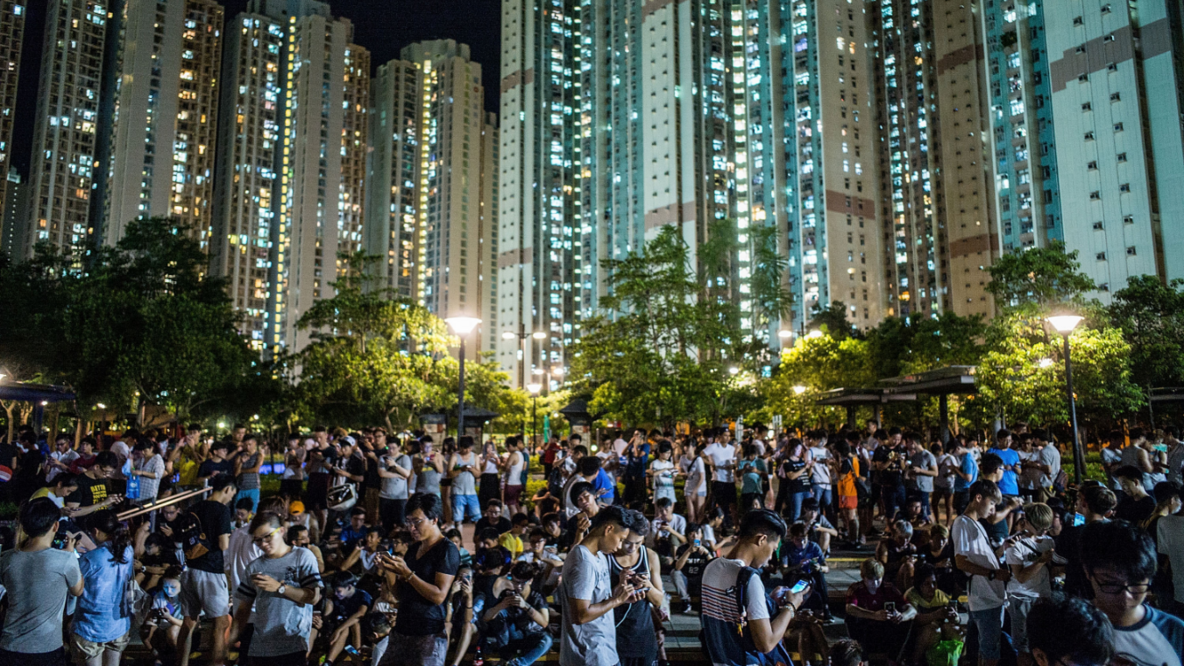 Thousands of young people are standing in a square, looking at their cell phones. The windows of the skyscrapers in the background are brightly lit. It is night.