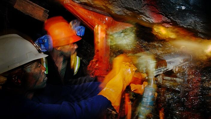 Two South African workers wearing helmets drill in a mine.