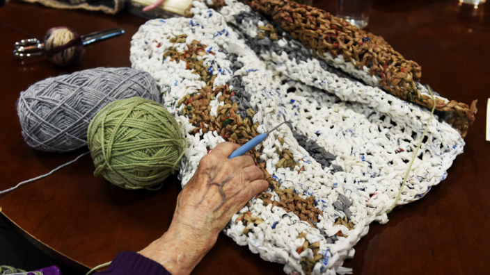 An older woman's hand with a crochet hook is lying on a crocheted iso-mat in white, brown and gray that is about half finished. Next to the mat lies a green and a gray "wool" ball. 