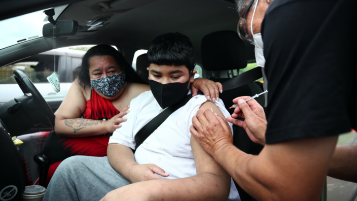 A boy is sitting in a car. His top is pushed up over his shoulder. A woman is sitting next to him. She has her arm around the boy's shoulder. A man is standing next to the open car door. The syringe in his right hand inoculates the boy. Everyone is wearing a health mask.