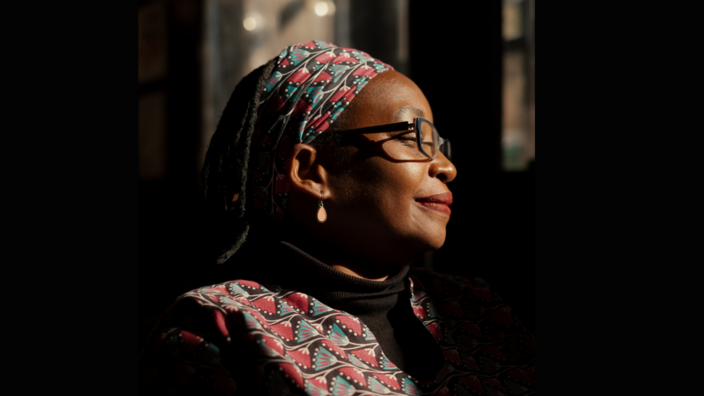 Portrait of a woman in a dark room, looking to the right. She is wearing a patterned headscarf and a matching collar. She is lit from the side and wears earrings and glasses. 