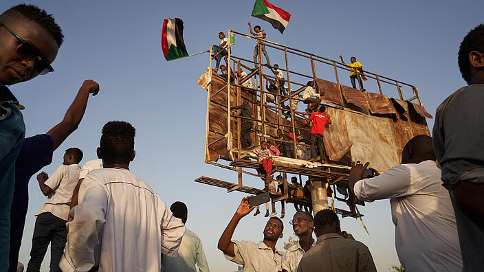 Several men are standing around outside. Several people are climbing on an improvised old scaffolding that towers into the sky. At the top, two young men are waving Sudanese flags