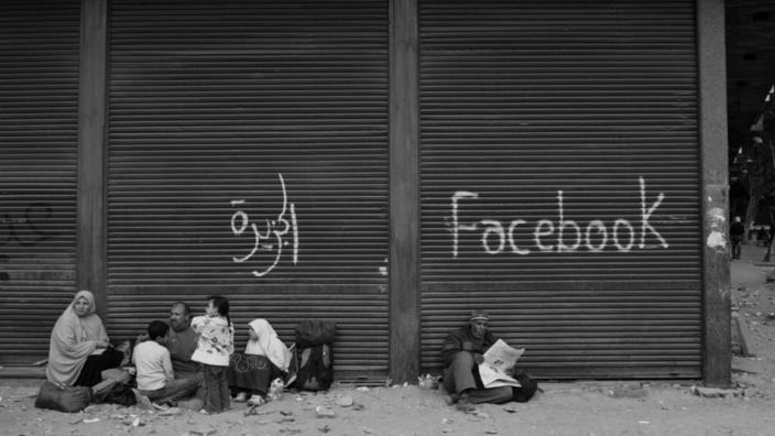 Eine arabische Familie sitzt auf dem Boden vor einem Gebäude mit geschlossenem Rollladen mit einer arabischen Aufschrift. Ein älterer Zeitung lesender Mann sitzt in unmittelbarer Nähe vor einem zweiten Rollladen mit der Aufschrift Facebook.