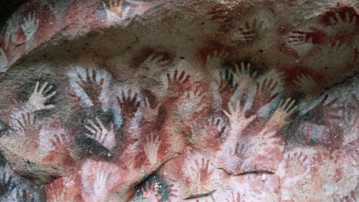 Auf einer grau, weiß, roten Höhlenwand sind viele verschiedene Hände abgebildet.