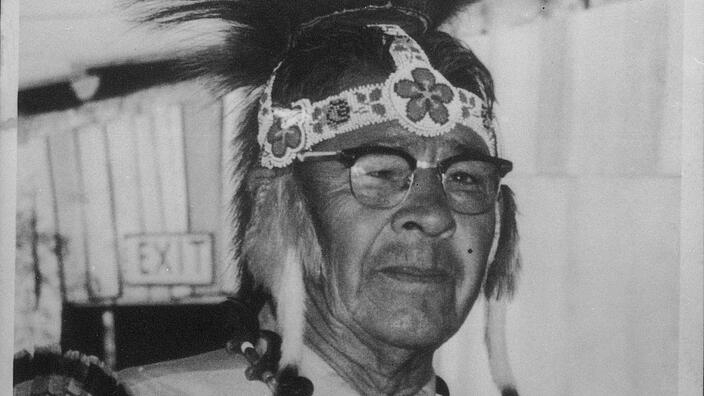A black and white portrait of an elderly indigenous man with a Native American headdress and necklace.