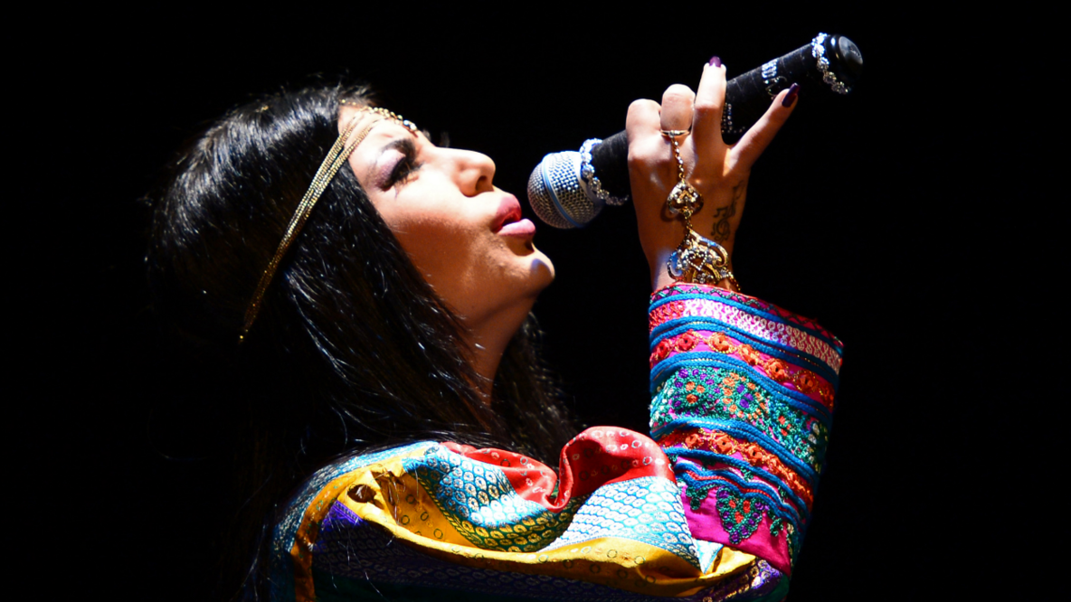 Afghan woman in profile. She has long dark hair and a headband. In her right hand she holds a microphone. The head slightly in the neck, the view diagonally upwards she sings. Her top is colorfully patterned in blue, yellow red, white.