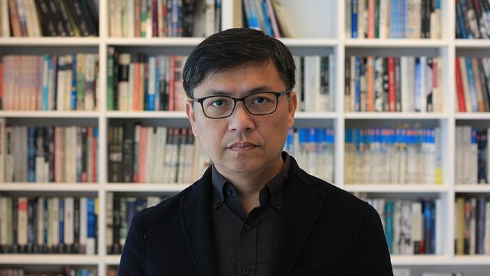 Wu Ming-Yi stands in front of a full white bookshelf. You can see him up to his chest. He wears a dark jacket and dark shirt. His look through the black metal glasses is serious.