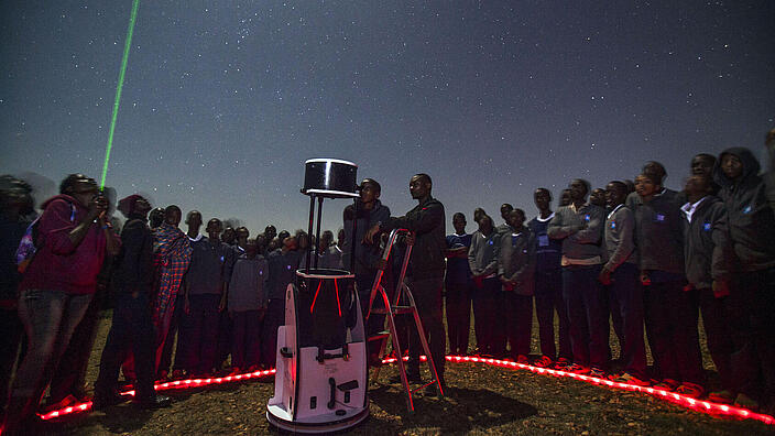 In front of a dark night sky with many stars, numerous young people are standing in a circle around a telescope that is pointed at the sky. Next to the telescope, a boy has climbed up a small ladder to look through the telescope