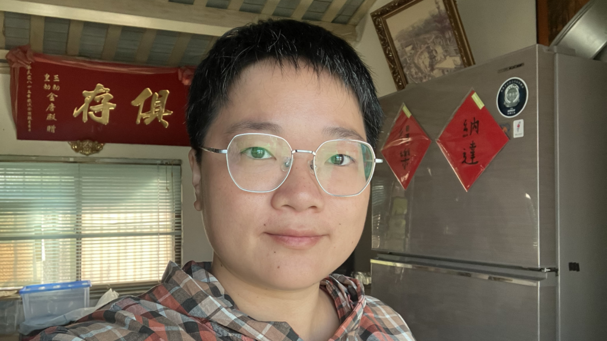 Chen Inzone ist eine junge Frau. Sie trägt kurzes Haar und eine Brille. Sie steht vor einem grauen Kühlschrank. Hinter ihr hängt ein rotes Banner mit chinesischen Schriftzeichen.