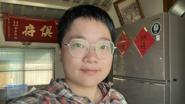 Chen Inzone ist eine junge Frau. Sie trägt kurzes Haar und eine Brille. Sie steht vor einem grauen Kühlschrank. Hinter ihr hängt ein rotes Banner mit chinesischen Schriftzeichen.