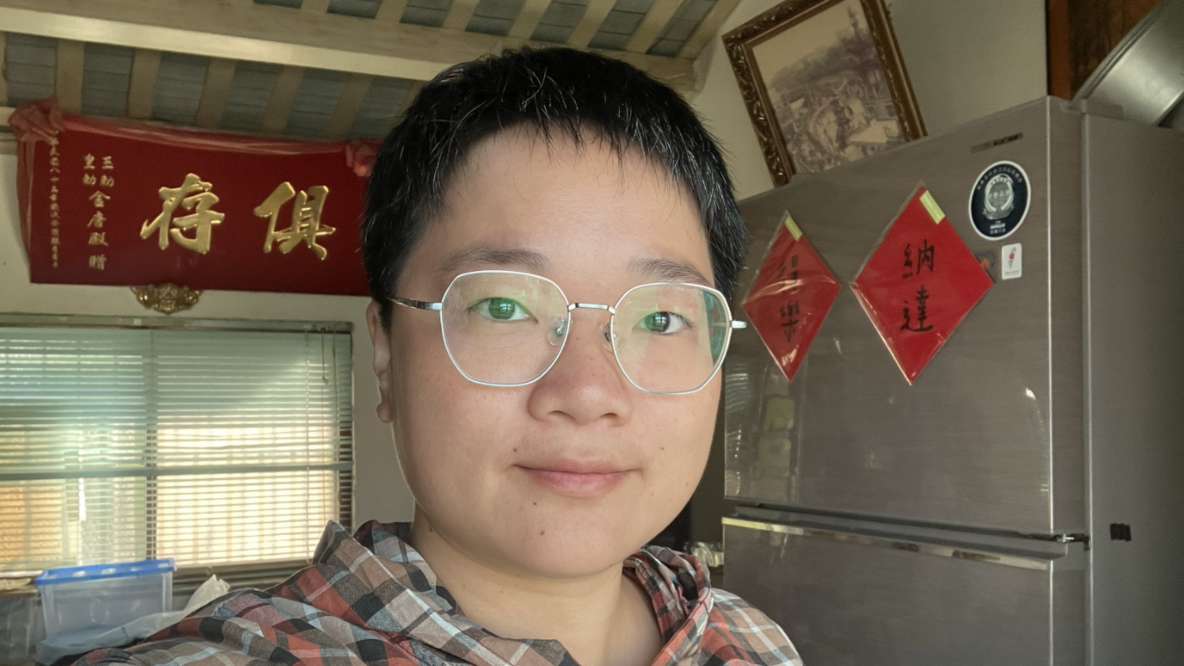 Chen Inzone is a young woman. She wears short hair and glasses. She is standing in front of a gray refrigerator. Behind her hangs a red banner with Chinese characters.
