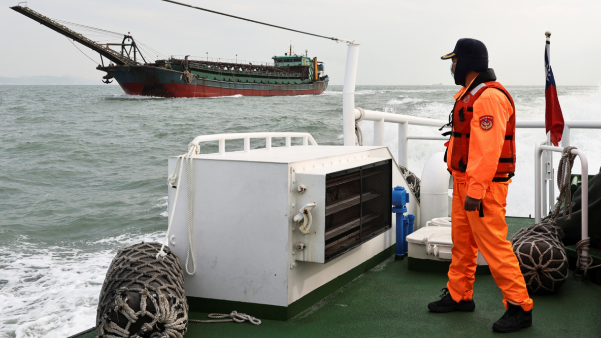 Ein Mann im leuchtend orangefarbenen Arbeitsanzug steht an Deck eines Schiffes und schaut zu einem Baggerschiff hinüber.