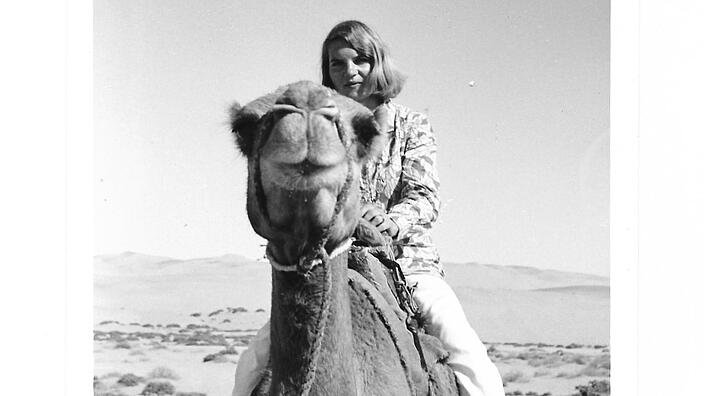 Black and white photo shows a young woman on a camel in a desert landscape. The woman is wearing long white trousers, a blouse and half-length blonde hair.