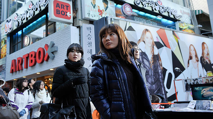 Two women in winter clothes can be seen in an urban environment. Behind them are the billboards of a large shopping centre. 