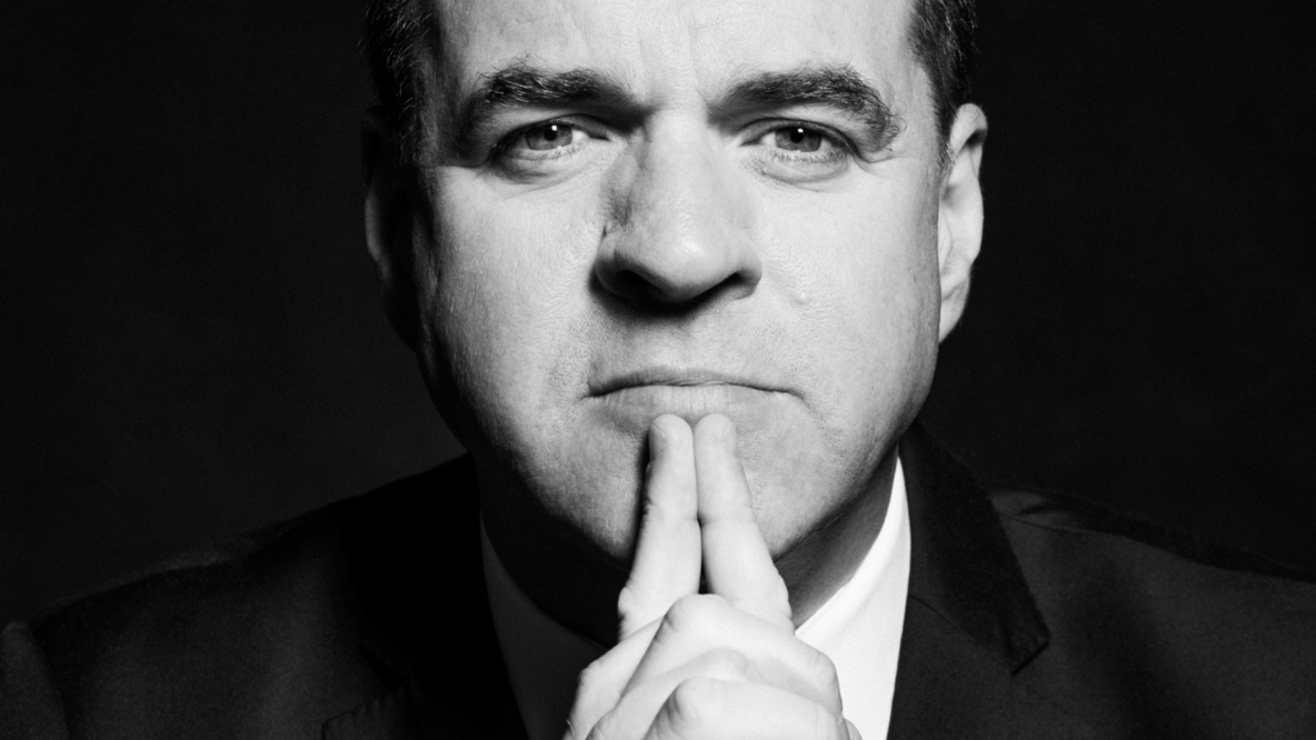 A black and white portrait of Niall Ferguson. He is looking directly into the camera. His hands are folded, the outstretched index fingers touch his lower lip.