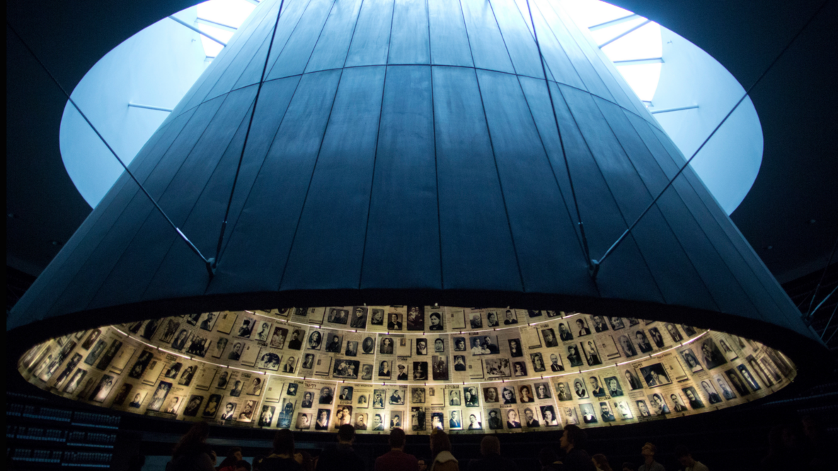 In a dark exhibition room, a circular memorial wall can be seen suspended from the ceiling. The inner wall is lined with many portraits and names.
