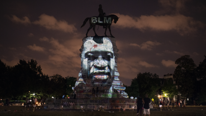 Eine Reiterstatue von General Robert E. Lee steht auf einem großen Sockel. Es ist Nacht. Auf den Sockel ist ein Gesicht projeziert. Es handelt sich um einen Afroamerikaner. Sein Name ist George Floyd.