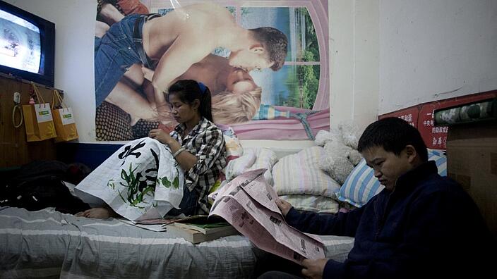 Das Bild zeigt ein junges chinesisches Ehepaar in einem engen Kellerraum, der wie ein Schlafzimmer eingerichtet ist. Die Frau sitzt auf dem Bett mit einer Handarbeit. Ihr Mann sitzt vor dem Bett und liest Zeitung.
