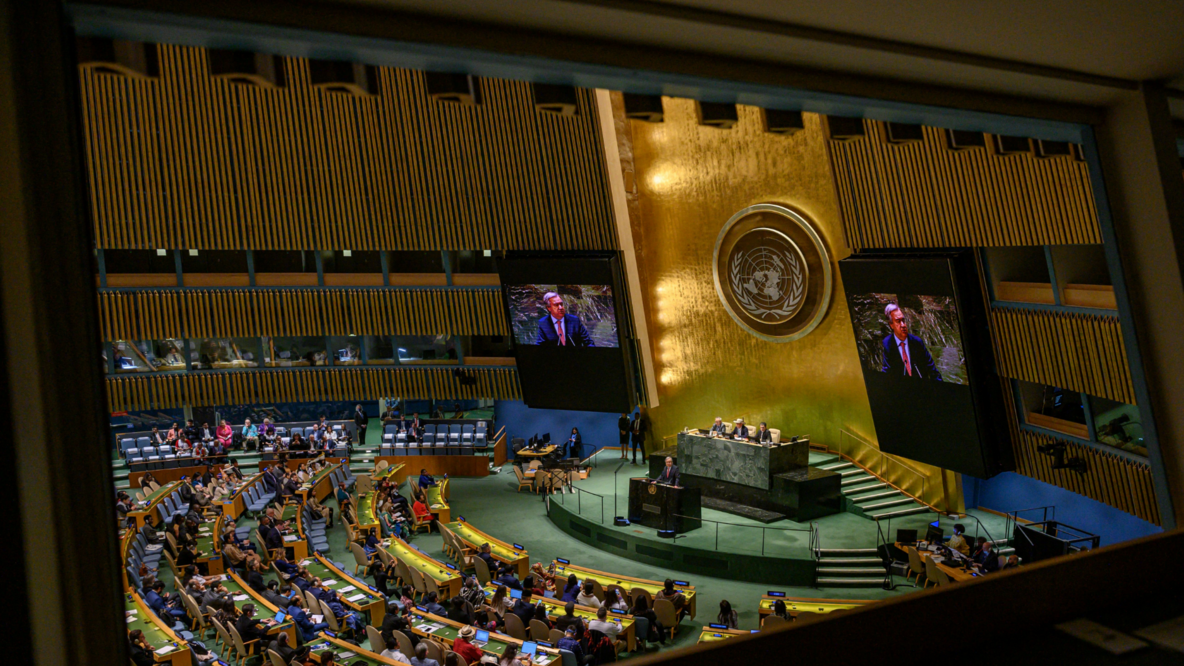 Through an observation window, the view goes into the assembly room of the United Nations. On the right side is the lectern, a central wall with the large UN logo, and two screens showing the speaker. On the left side, the audience sits in rows of seats 