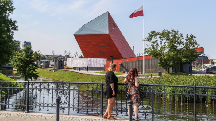 Two young women are standing on the railing of a bridge. They are looking into the water. In the background are three modern buildings in red and orange, with the Polish flag in front of them.
