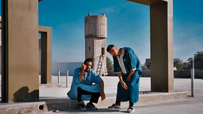 In a heavily cemented setting against a very blue sky, two young men pose in equally blue kaftans with white embroidery, wearing sneakers