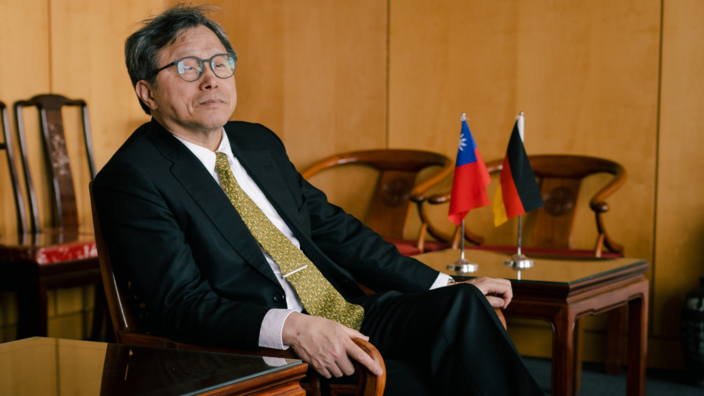 Shieh Jhy-Wey is sitting on a chair in his office. He wears glasses, a dark suit and a yellow and green patterned tie. On the table behind him are miniature flags of Germany and Taiwan.