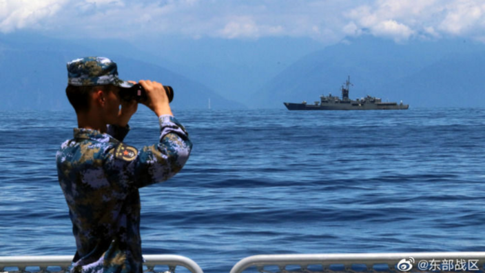 Ein Soldat in chinesischer Uniform schaut durch ein Fernglas. Im Hintergrund ist ein großes Schiff zu sehen, außerdem Berge.