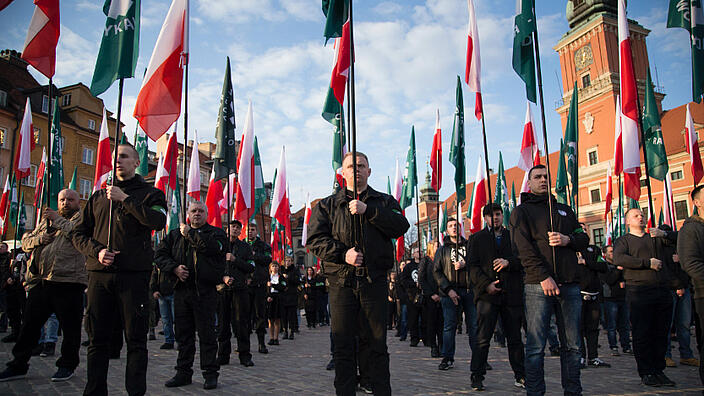 Viele Menschen stehen mit gleichmäßigem Abstand und rot/weiß und grünen Flaggen auf einem Platz.