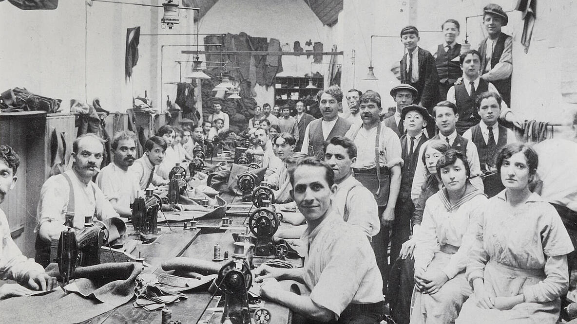 Das Schwarz-Weiß-Foto zeigt den Blick in eine große Manufaktur-Werkstatt. An einem langen Tisch sitzen zahlreiche Männer an Nähmaschinen, entlang der Wände sind weitere Männer und Frauen aufgereiht. Alle schauen für das Gruppenporträt in die Kamera