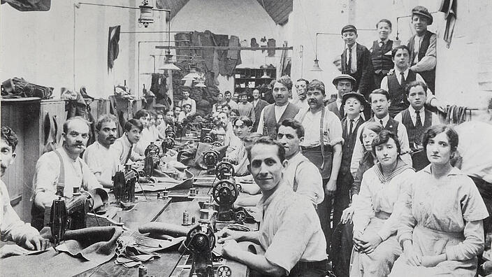 Das Schwarz-Weiß-Foto zeigt den Blick in eine große Manufaktur-Werkstatt. An einem langen Tisch sitzen zahlreiche Männer an Nähmaschinen, entlang der Wände sind weitere Männer und Frauen aufgereiht. Alle schauen für das Gruppenporträt in die Kamera