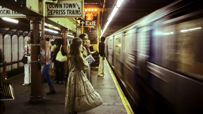Mehrere Menschen stehen an einer Subway Station in New York City. Eine U-Bahn fährt gerade ein. Zur Orientierung wurden verschiedene Schilder angebracht. (Exit, Down Town Express Trains) 