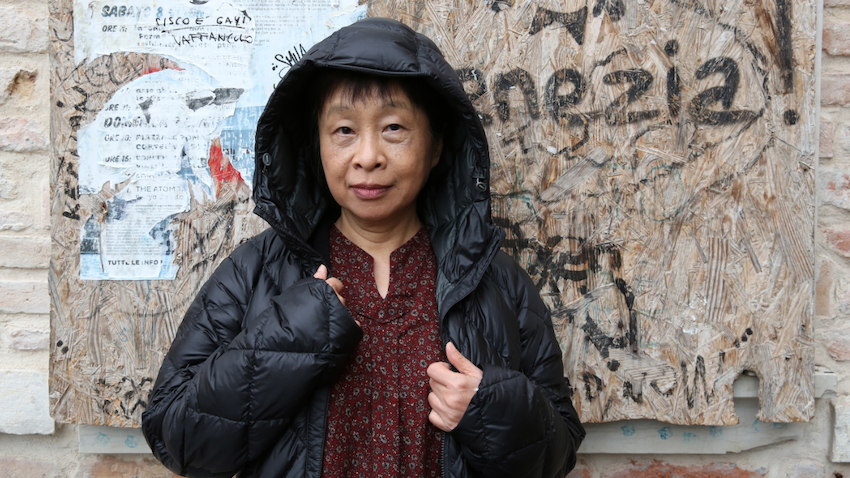 Eine Frau mittleren Alters steht in einem Hauseingang. Die Wände bestehen aus rohen Backsteinen mit abgerissenen alten Postern. Die Schriftstellerin trägt eine schwarze Jacke mit aufgezogener Kapuze und blickt in die Kamera