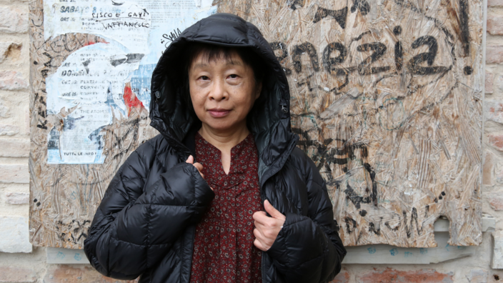 A middle-aged woman stands in a nondescript doorway. The walls are made of rough bricks with torn old posters. The writer wears a black jacket with the hood up and looks into the camera