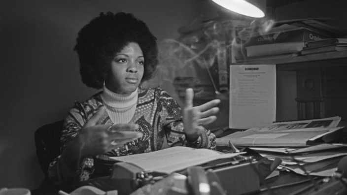 The photo is black and white. Young Margaret Busby is sitting behind her typewriter, gesturing with her hands. Next to her in the ashtray, smoke rises from a cigarette.