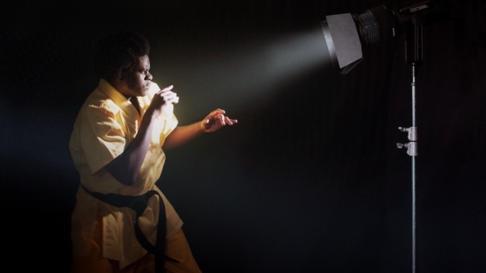 Black background. In the lower left corner is a black man in a light yellow almost white judo suit. The man has his head tucked in, his arms are in front of his body in a defensive posture. Opposite him is a luminous spotlight, the light of which is directed at the man. 