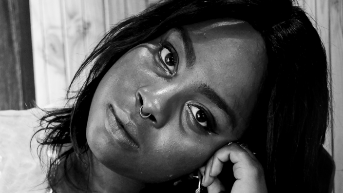 Ein schwarz weißes Fotoporträt von Binwe Adebayo. Sie hat dunkle Haare. Sie stützt ihren geneigten Kopf mit der linken Hand.