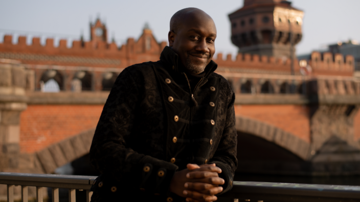 Musa Okwonga leans against a railing, he smiles into the camera. In the background you can see the bridge Oberbaumbrücke in Berlin.