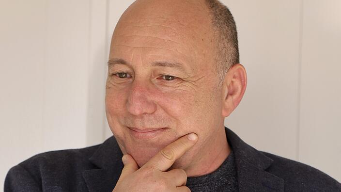 A portrait of Ronen Kadushin. He wears his few hairs very short. The index finger and thumb of his right hand rest on his chin. He smiles and looks past the camera on the right. 