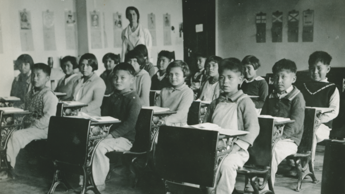 In einem Klassenraum sitzen Kinder etwa im Alter von 7 Jahren hinter ihren Pulten. Im Hintergrund steht eine Lehrperson