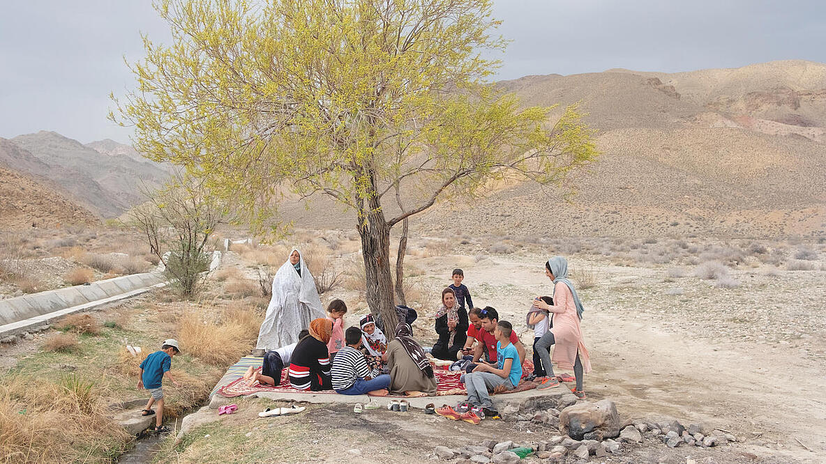 On a stony plateau, a group of people are sitting on several blankets under a tree. There are women, men and children of different ages. A little to the side, a boy dips his foot in a small stream. Mountains can be seen in the background. 