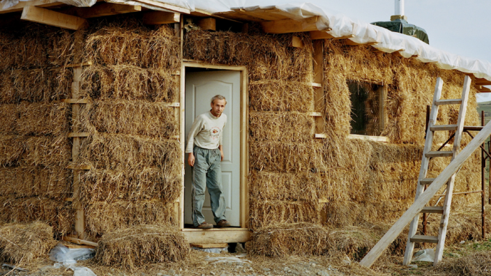 The outer wall of a one-story house is insulated with straw, there is no external facade. A ladder leans against the house, a middle-aged man steps out of the open door onto the dirt floor. He wears jeans and a long-sleeved T-shirt.