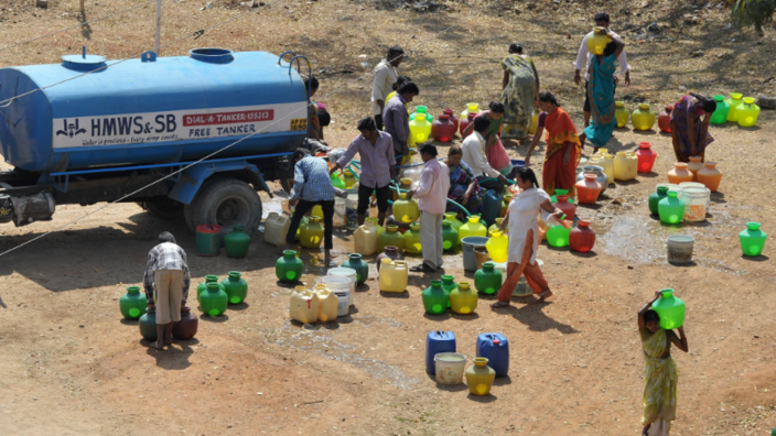 People with many colorful canisters stand around a blue tanker truck to fetch water.