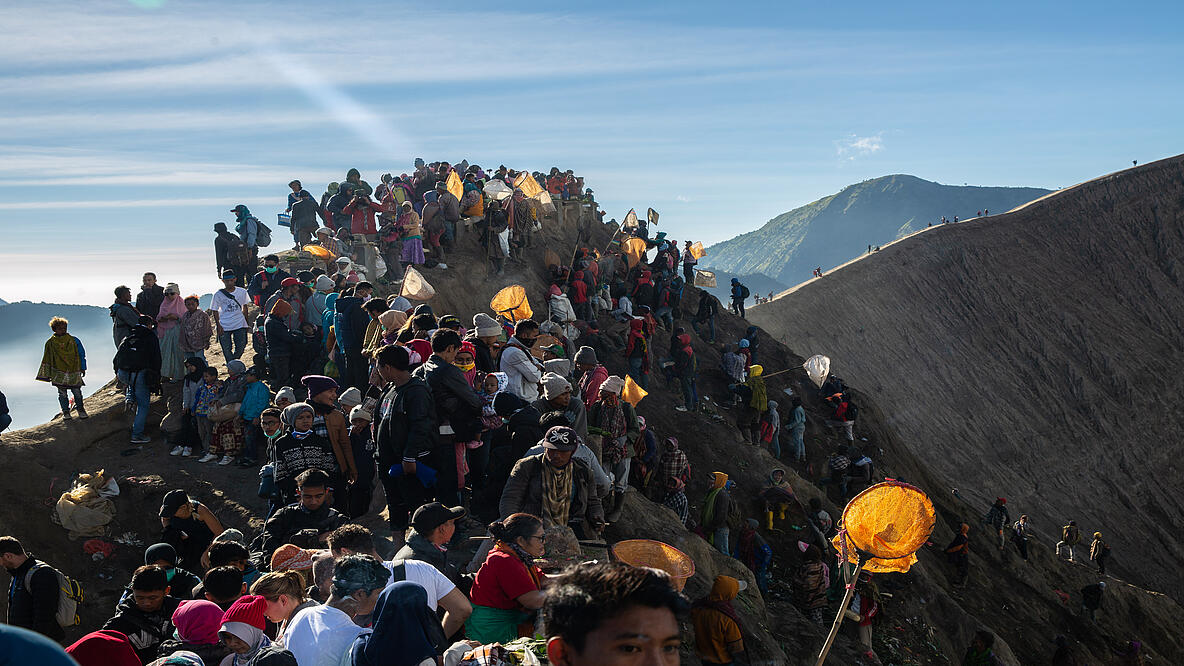 Auf einem Berggipfel stehen und sitzen viele Menschen. Weitere Menschen befinden sich noch auf dem Weg zum Gipfel. 